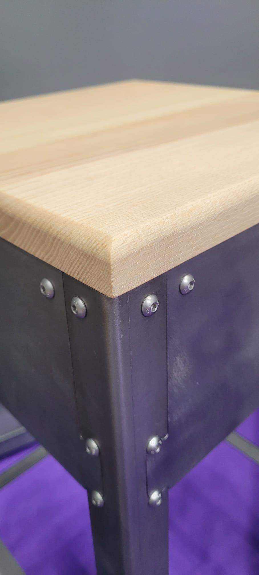 Gros plan sur un tabouret design, montrant l'assemblage d'une assise en frêne massif du Cantal sur une structure en acier brossé avec visserie inox.