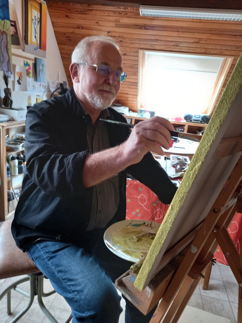 L'artiste Francis Heintz assis devant son chevalet en train de peindre. Habits noir et Bleu; Fond: déco atelier en brun clair et blanc.