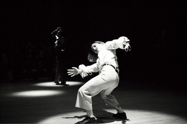 Photo d'une personne habillée en blanc, avec un pantalon blanc, une ceinture et une chemise bouffante. Cette personne a les cheveux longs et une moustache, ses bras s'agitent dans une posture dramatique, comme dansant, en transe. Les éclairages sont des éclairages scéniques de théâtre dans une atmosphère sombre. Une silhouette noire apparaît en fond quelques mètres derrière.