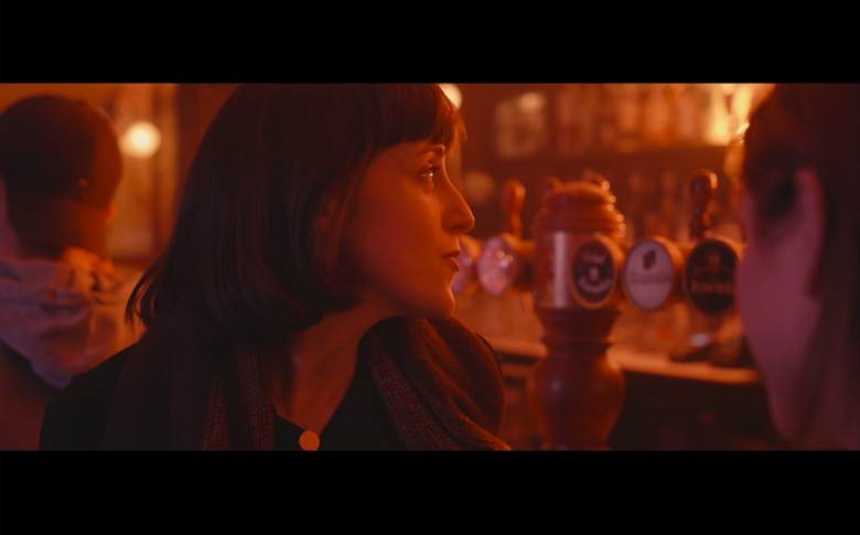 Une femme blanche est de profil, dans un bar. Elle a les cheveux longs et regarde derrière elle. Au premier plan, on discerne le visage d'une autre femme blanche qui semble aussi avoir environ 25 ans.