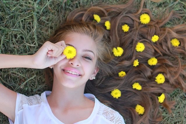 Cette image est une photo d'une adolescente allongée dans l'herbe, ses longs cheveux châtains étendus sur le côté gauche de son visage. Des fleurs jaunes sont éparpillées sur ses cheveux. Elle en tient une sur son oeil droit, le sourire aux lèvres.