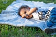 Cette image est une photo d'un petit garçon d'environ 3-4 ans, allongé sur une couverture dans l'herbe, calme. Il semble vouloir toucher l'herbe avec ses doigts. Cet enfant inspire la tranquilité, le repos, l'exploration.