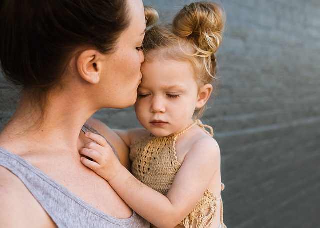 Cette image représente une petite fille dans les bras de sa maman. Une main de la petite fille est posée dans le cou de la maman qui l'embrasse sur le front.