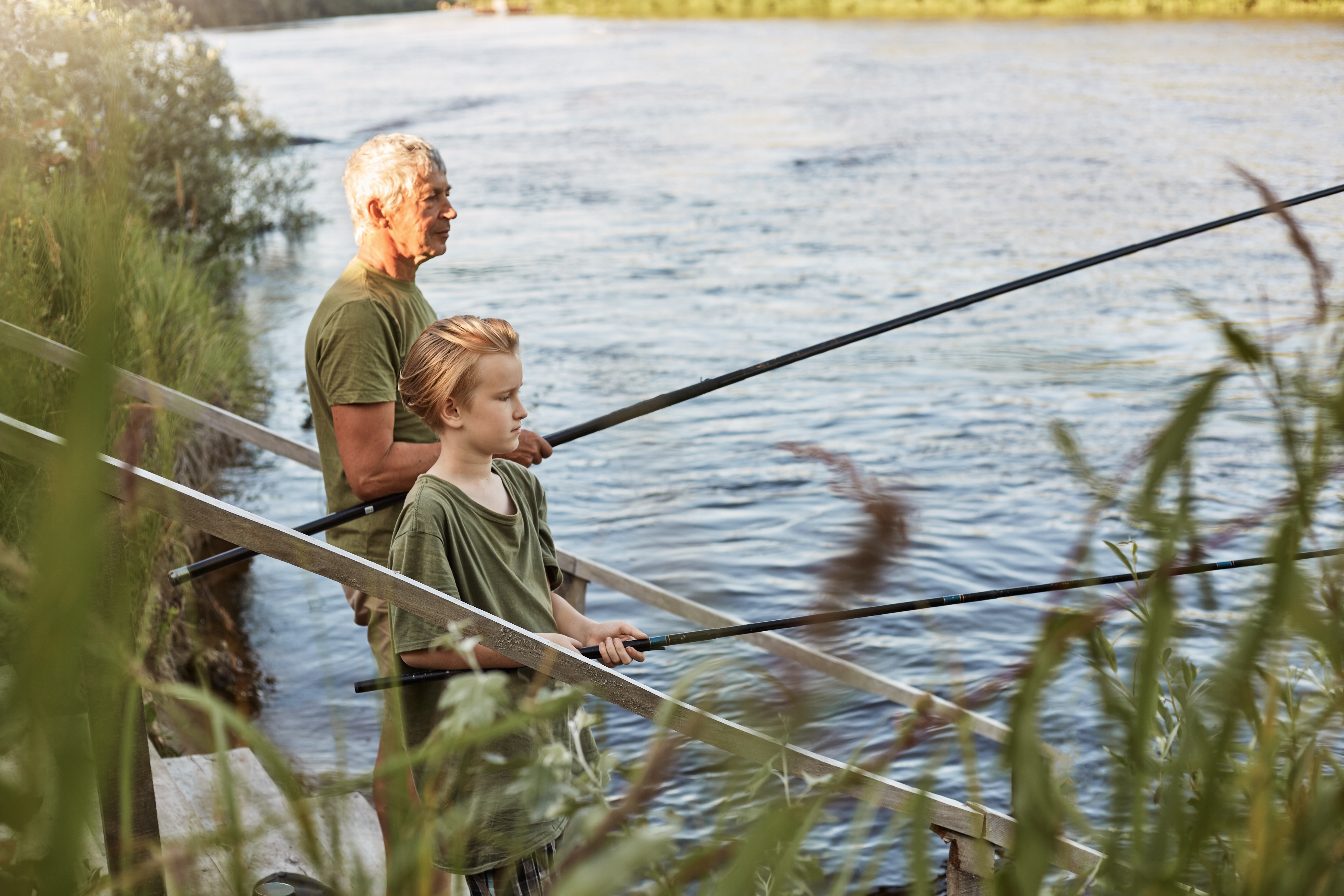 European-grey-haired-mature-father-with-son-outdoors-fishing-by-lake-river-standing-near-water-with-fishing-rods-hands-dress-casually-enjoying-hobby-nature