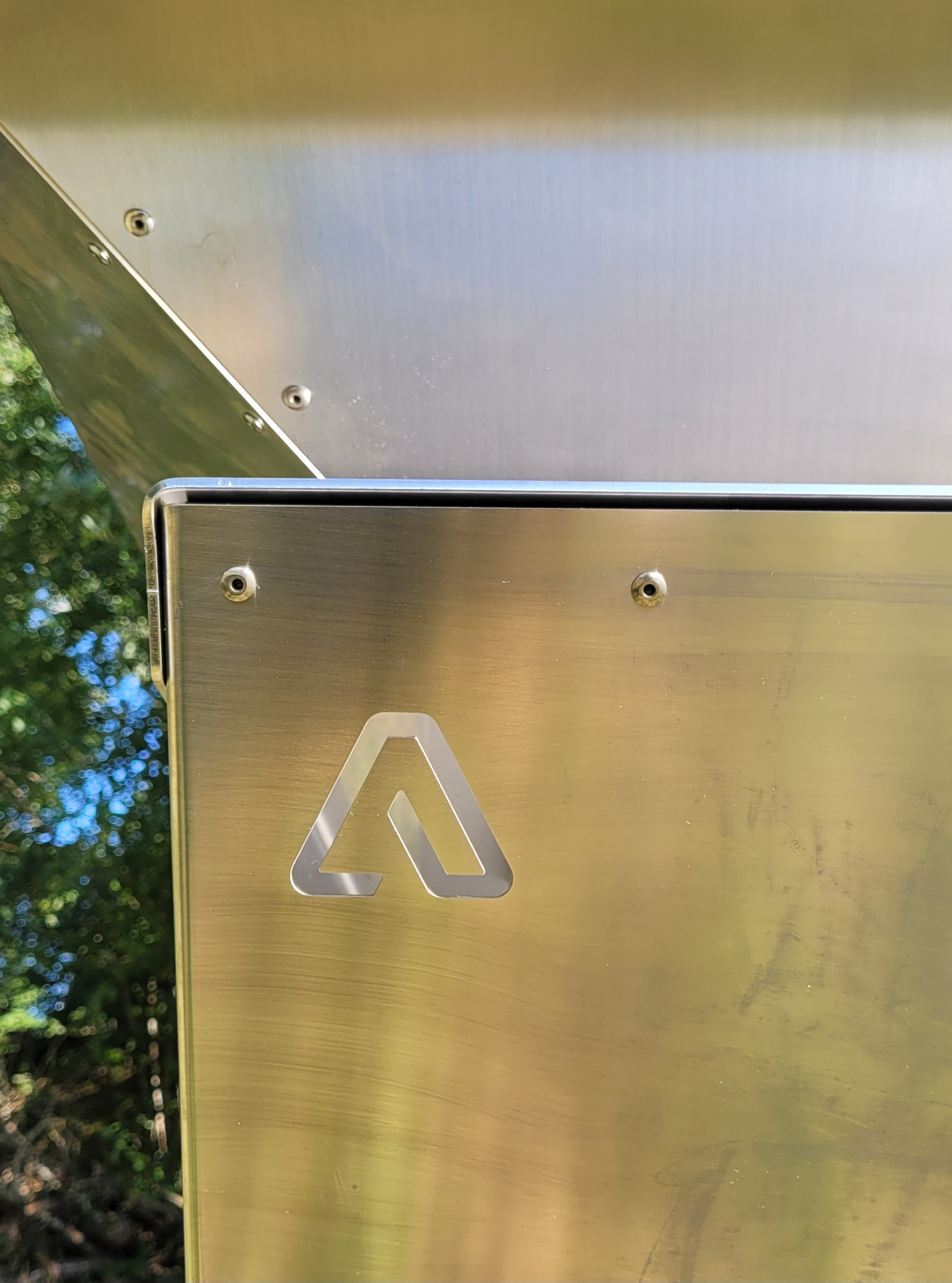 Vue détaillée de la finition d'un brasero en inox brossé, montrant un assemblage par rivets et le logo 'A' de l'Atelier Créateur découpé au laser