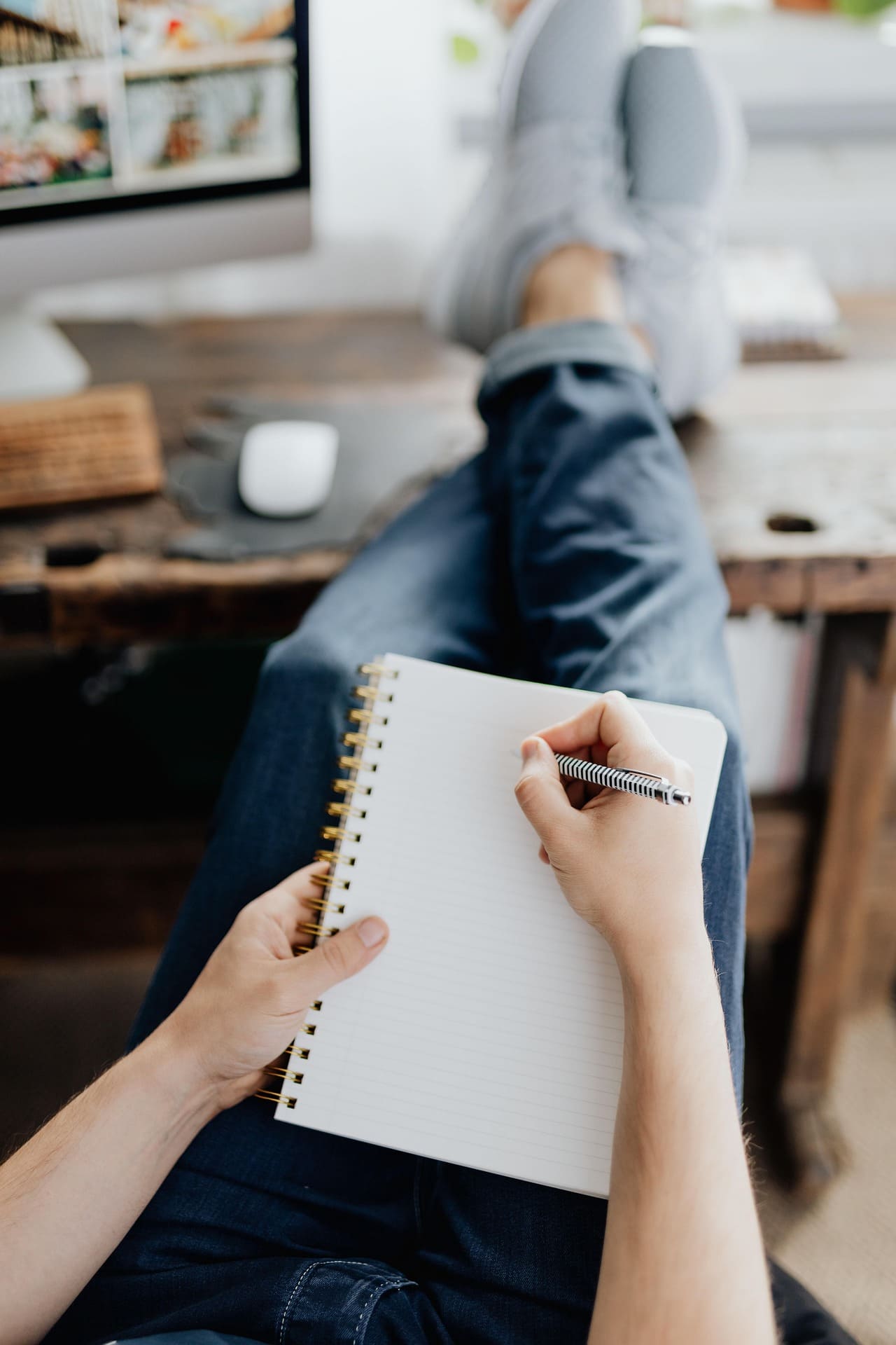 Cette image représente une personne installée les pieds croisés sur un bureau en bois. La personne écrit sur un cahier posé sur ses genoux.