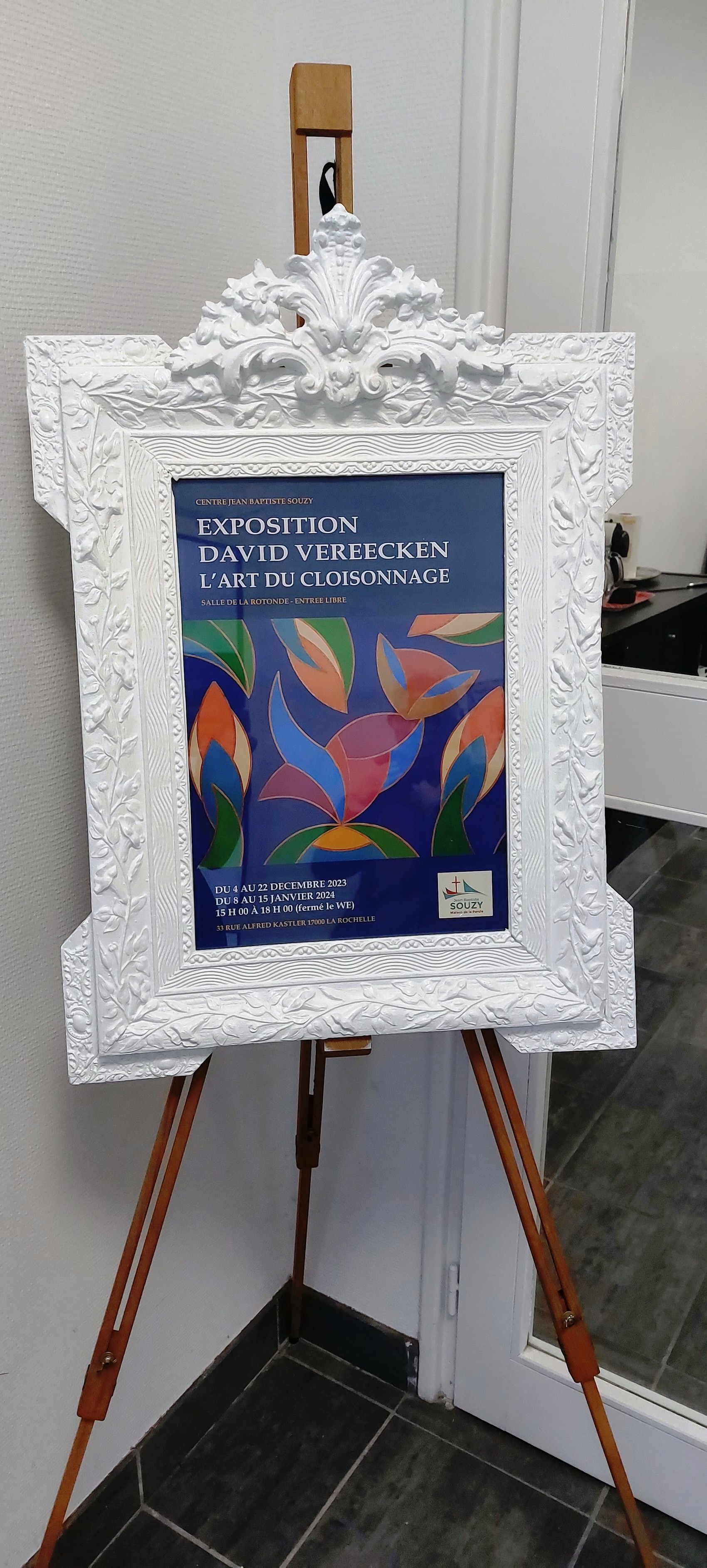 Affiche de l'exposition de David Vereecken présentée dans un cadre sculpté blanc sur un trépied à l'entrée de la galerie.
